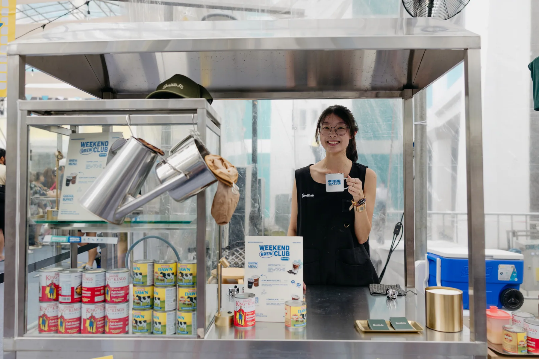 Coffee stall team member holding a Weekend Brew Club enamel mug beside the brew counter